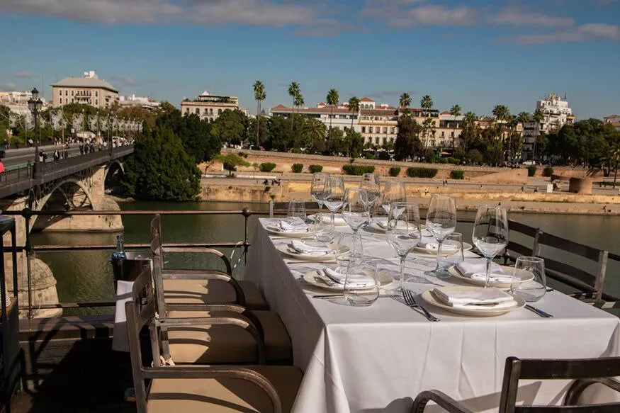 A table laid with white linen and glasses on a terrace above the Guadalquivir, the Triana bridge to the left and palms along the far bank