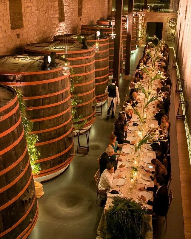 A long candlelit table set with white flowers in a stone-walled cellar, beside a row of giant oak vats bound with red hoops, guests seated along both sides and a waiter in a long apron