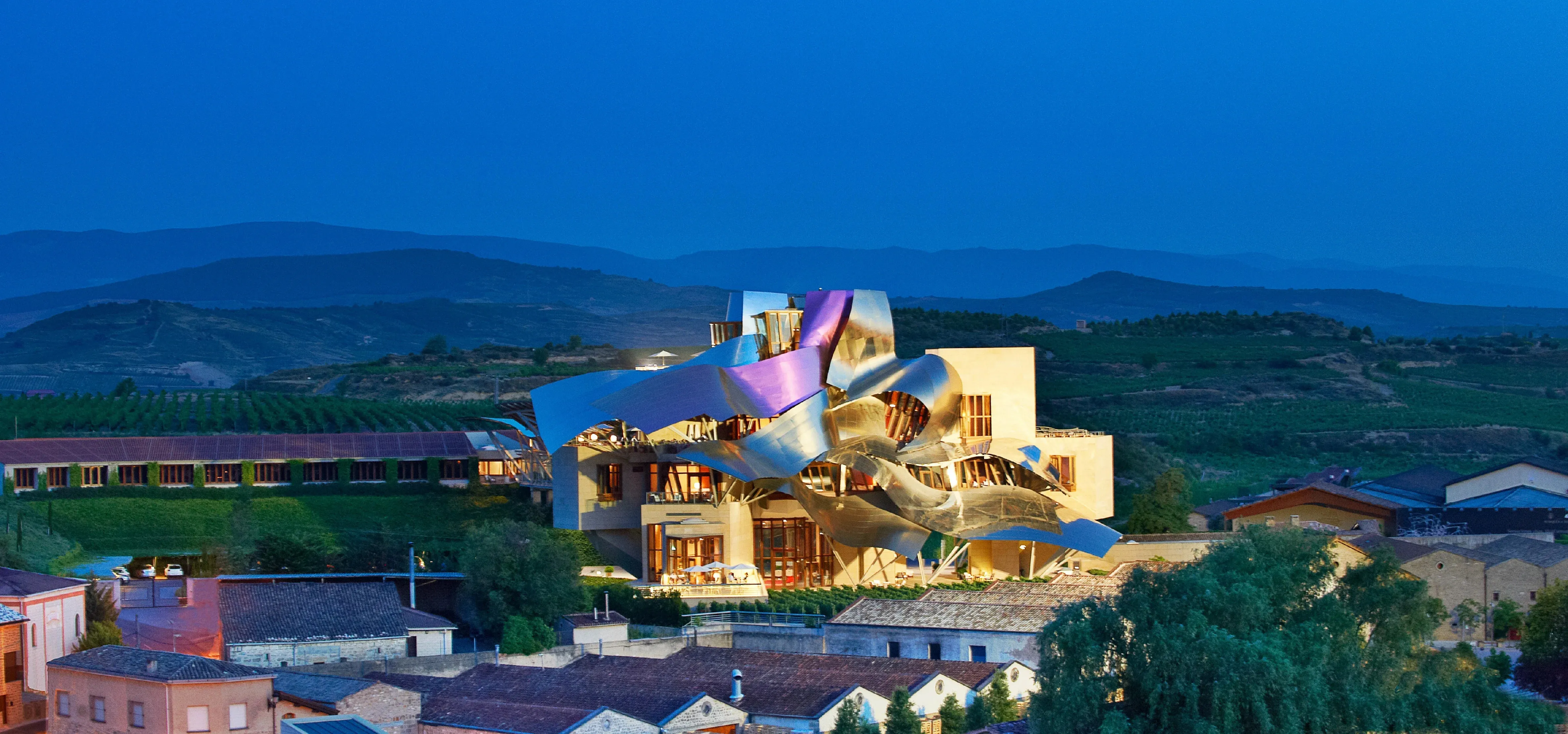 Hotel Marqués de Riscal at dusk, its rippling roofs of pink, gold and silver metal lit above the tiled roofs of Elciego, with vineyards and a line of low hills beyond under a deep blue sky