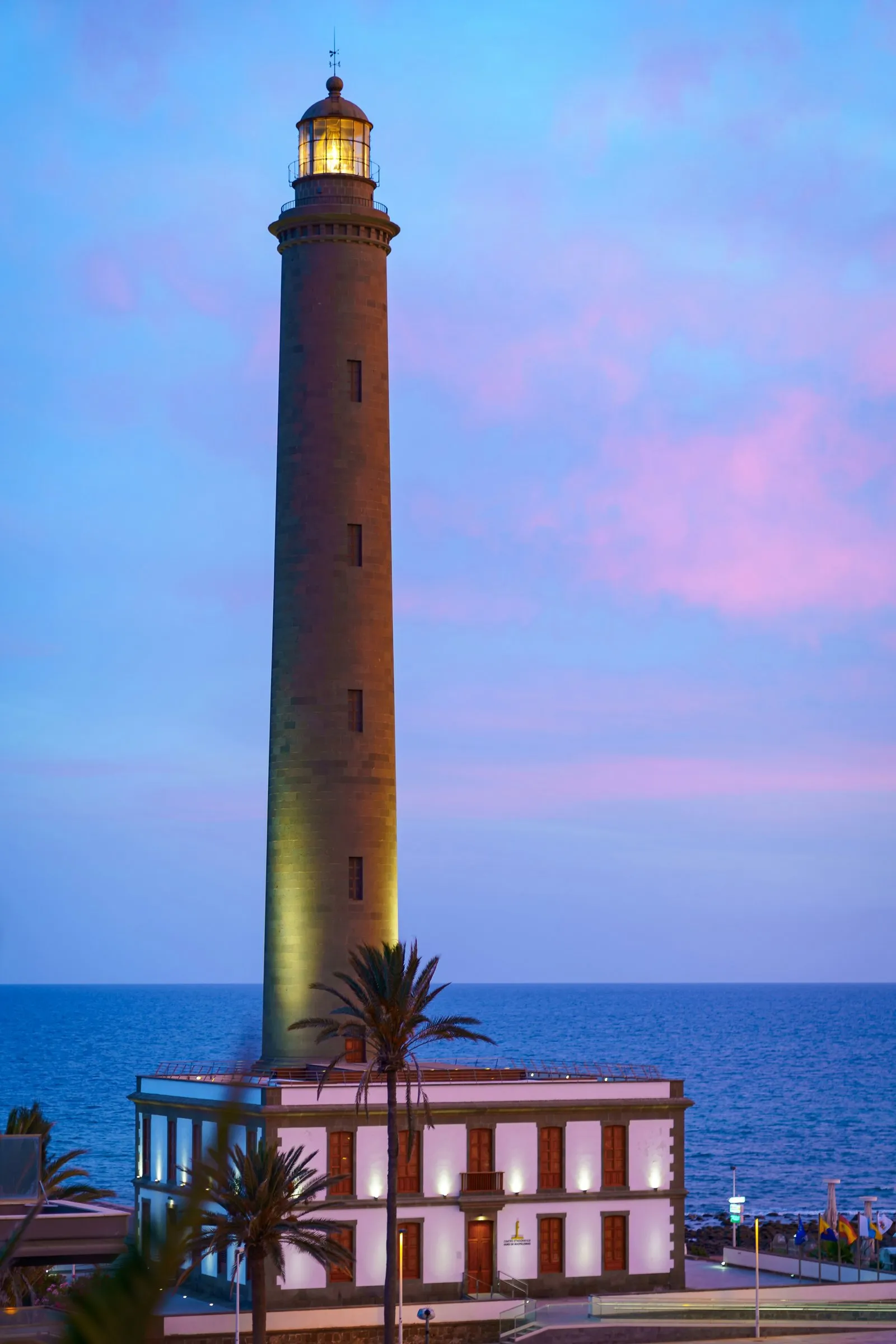 A tall stone lighthouse lit at dusk above a white two-storey house with brown shutters, palms in front and the sea behind under a pink sky