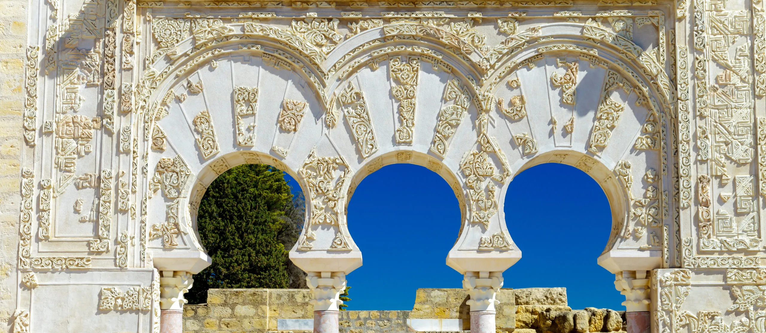 Three horseshoe arches of carved white stone at Medina Azahara, their voussoirs and frame covered in carved plant and geometric ornament, on pink marble columns, with blue sky and a cypress seen through them and ruined walls below