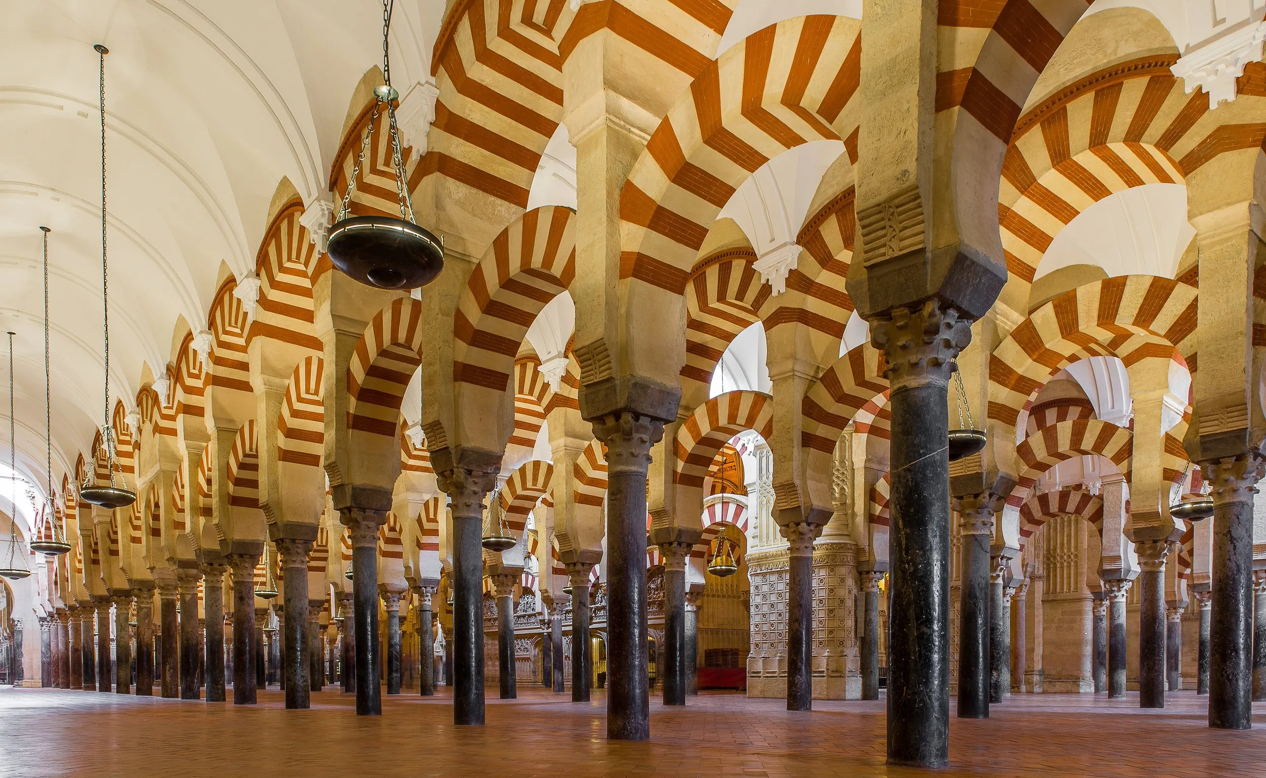 Rows of columns in the prayer hall of the Mezquita-Catedral, carrying two tiers of horseshoe arches striped in red brick and pale stone, bronze lamps hanging on long chains beneath a white vaulted ceiling on the left