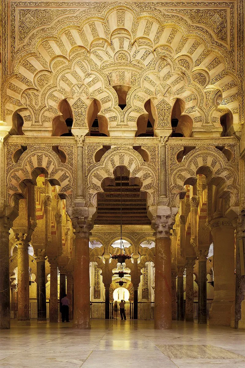 Interlaced, many-lobed arches of carved cream stone above marble columns inside the Mezquita-Catedral, further arcades receding into lamplight beyond, two small figures far off