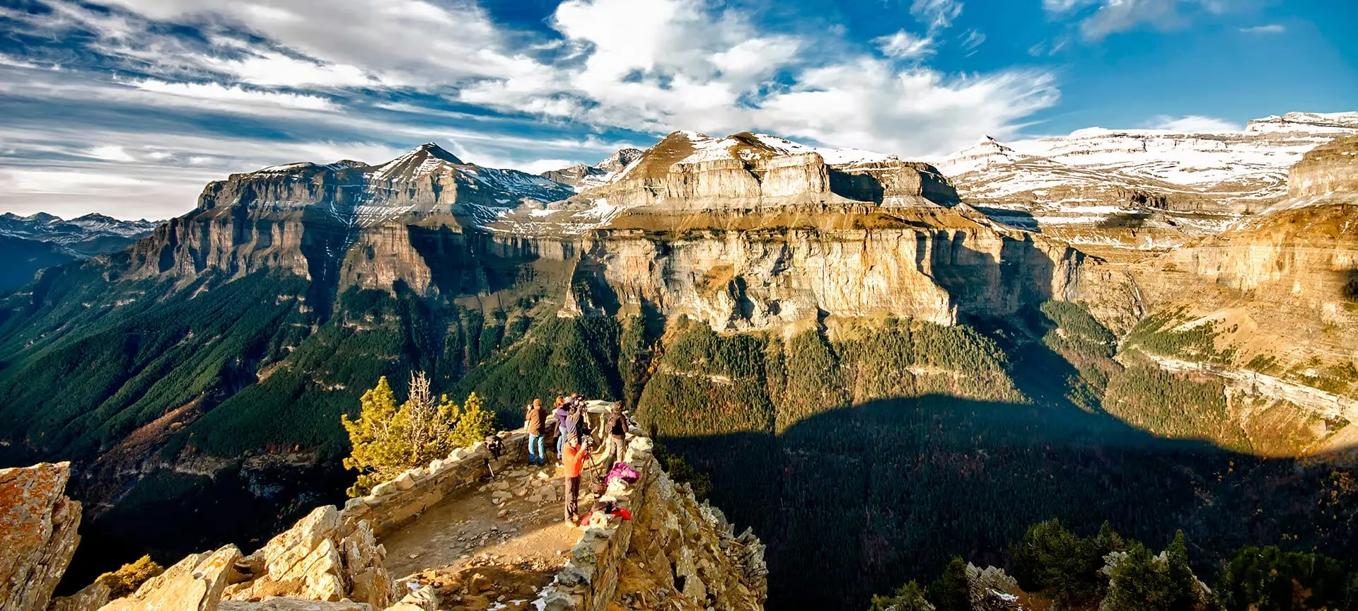 Walkers and photographers at a stone-walled viewpoint on a rocky spur, high above a forested canyon whose limestone walls and snow-dusted summits are lit by low sun under streaked cloud