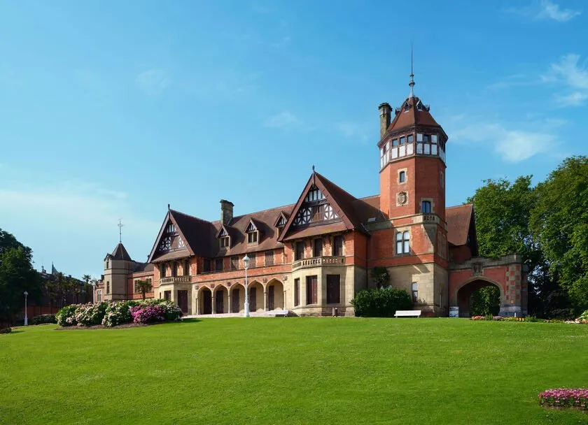 A red-brick mansion in the English country-house style, with steep gables, half-timbered dormers and a corner tower capped by a spire, above a wide sloping lawn with beds of pink hydrangeas, under a blue sky