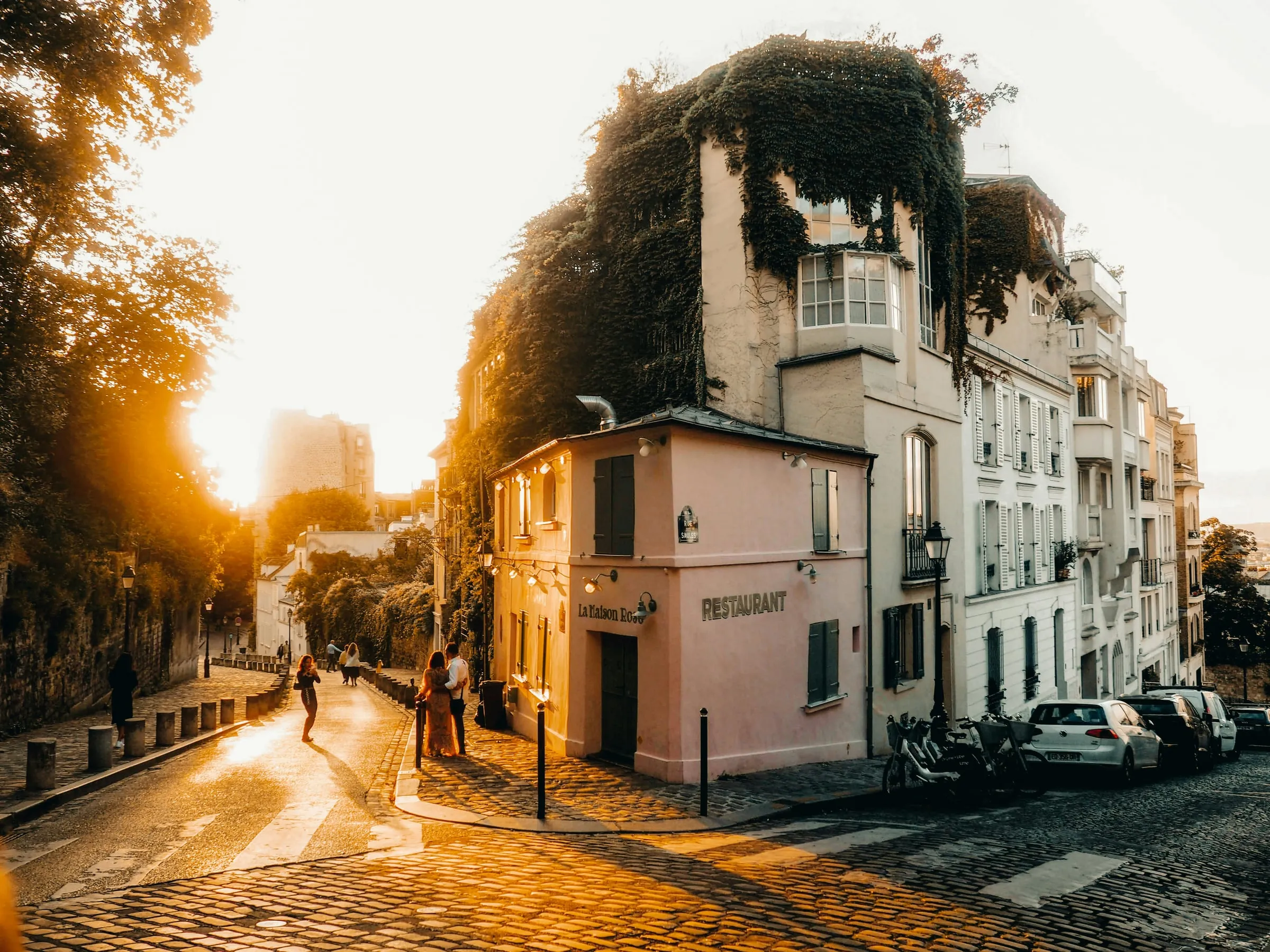 A cobbled corner of Montmartre at sunset: an ivy-covered pink house with green shutters where two sloping streets meet, white houses with grey shutters beyond it, and low golden light coming through the trees on the left