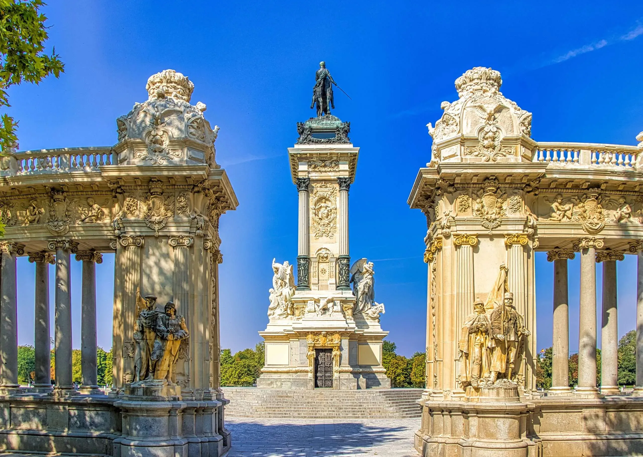 An equestrian statue on a tall column, framed between two curved stone colonnades with sculpted groups at their ends, under a deep blue sky