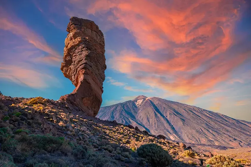 Roque Cinchado, a weathered column of rock, standing on a scrubby slope against a pink and orange sunset sky, with the cone of Teide behind it