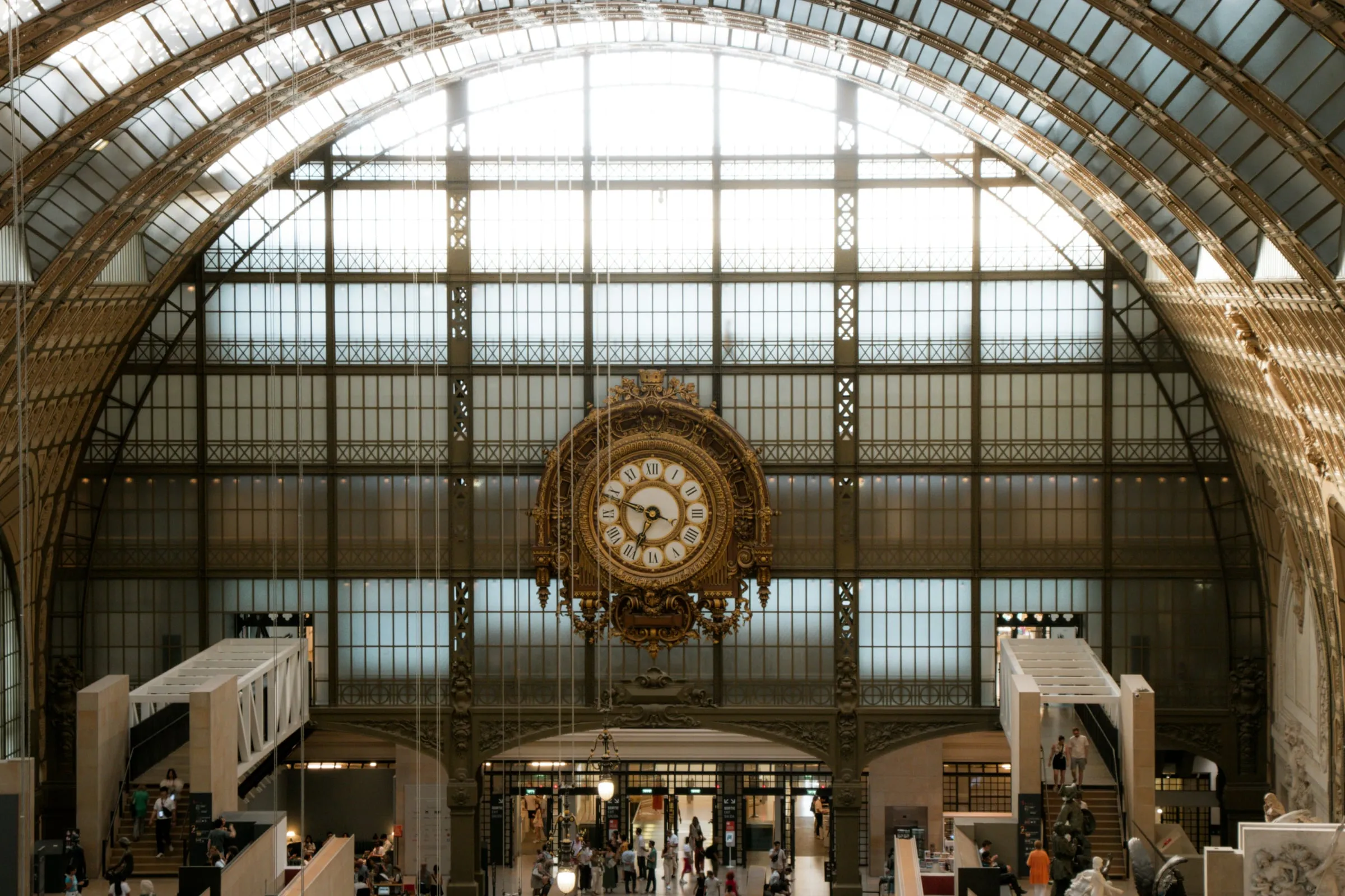 The great hall of the Musée d'Orsay under its glass roof: the gilded station clock set in the end wall, the iron arches of the old train shed above it, and visitors small on the floor below