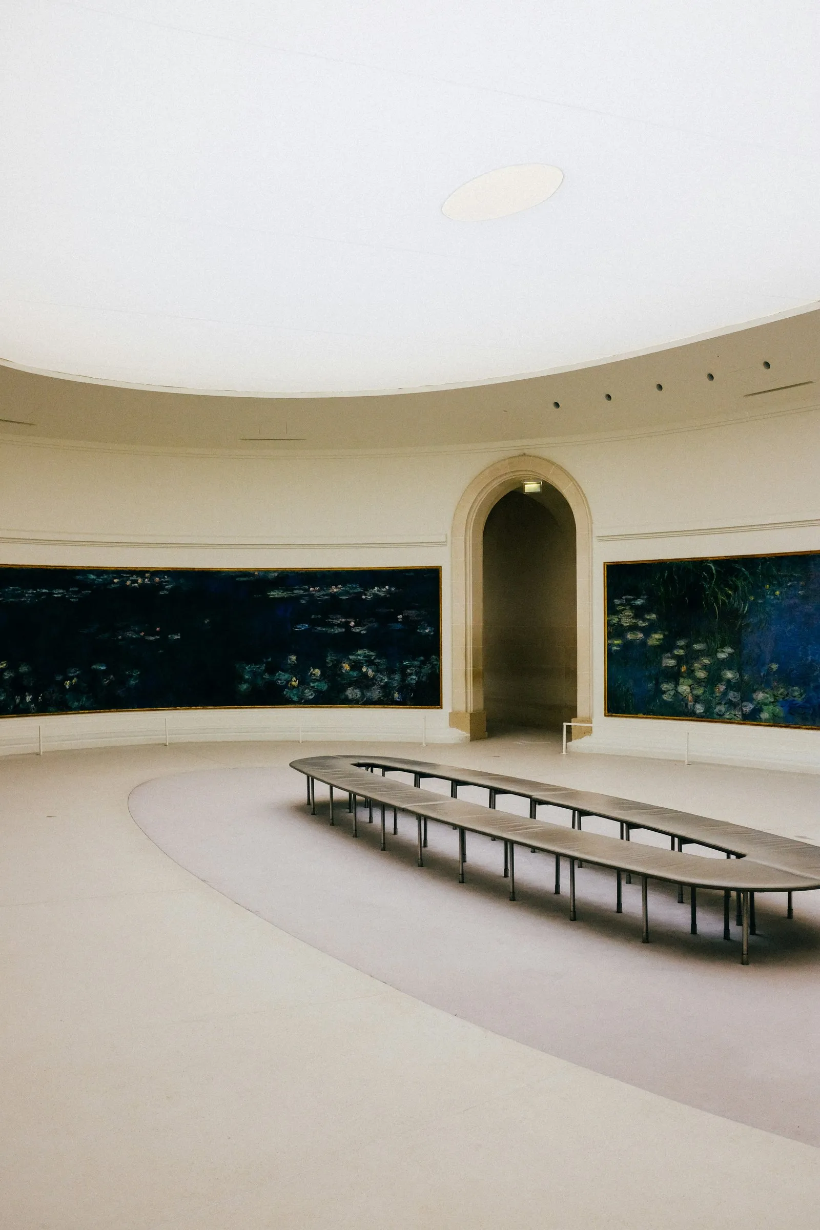 One of the two oval Nymphéas rooms of the Musée de l'Orangerie, empty: the curved water-lily panels in their gilt frames along the white walls, the arched opening through to the second oval room, and the long oval bench on the pale floor