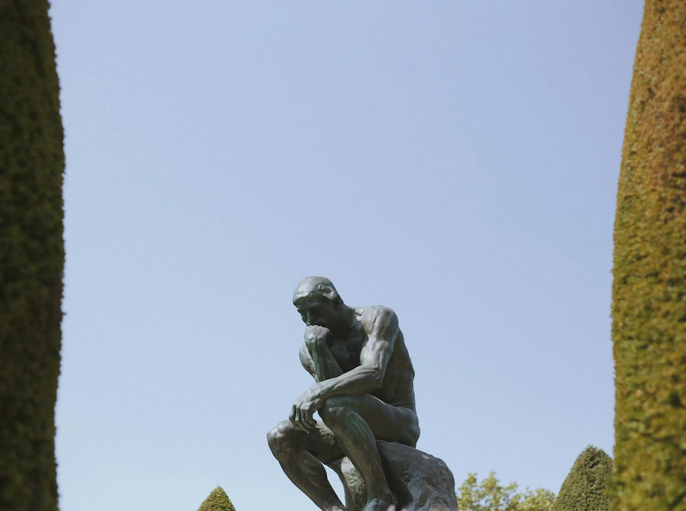 Rodin's The Thinker in bronze, seated with his chin on his hand, against a clear pale sky between two tall clipped yew cones in the museum's garden
