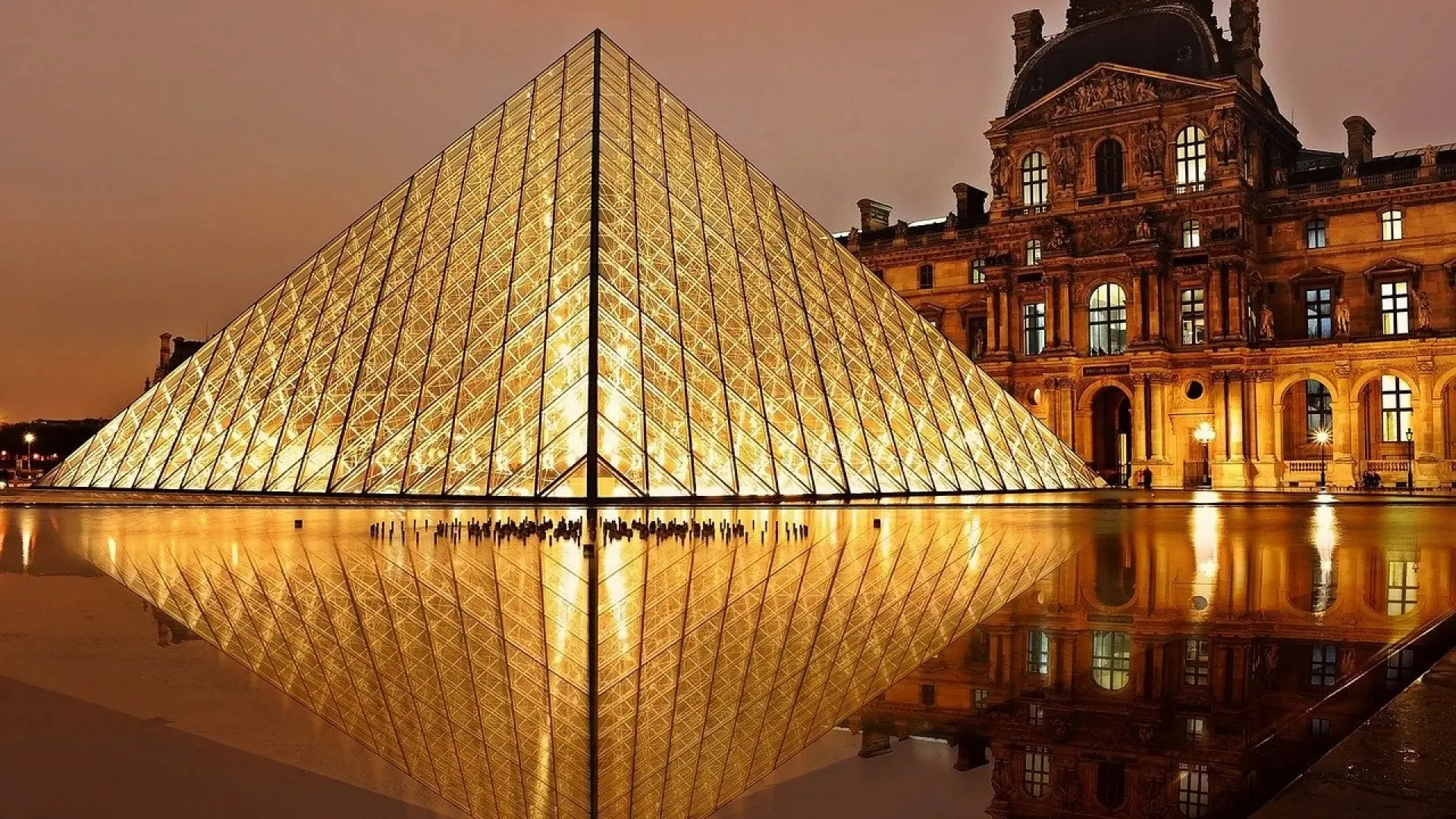 The glass pyramid of the Louvre lit gold at night and mirrored in the still water of a pool, with the lit stone façade of a palace pavilion on the right under a dusky sky
