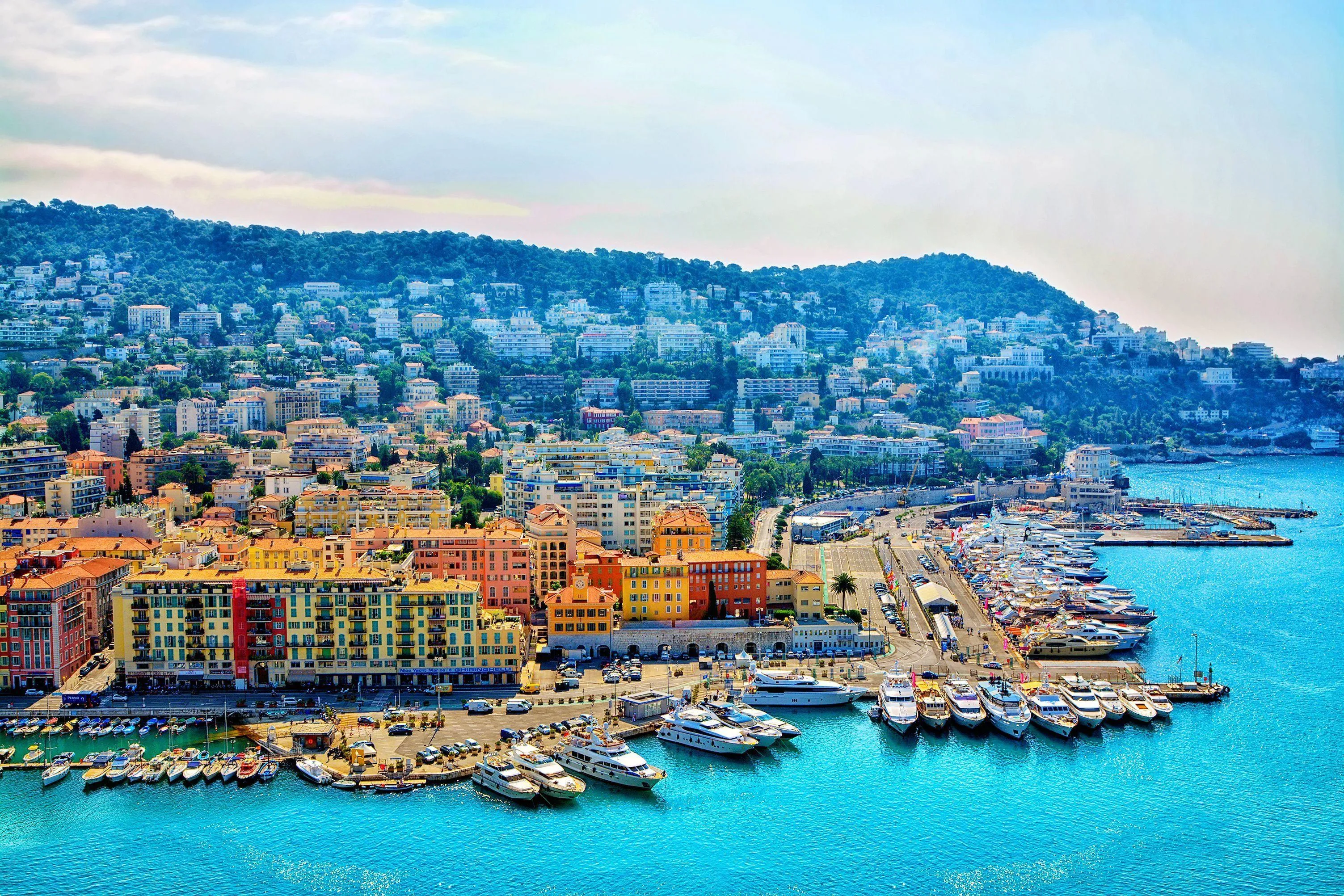 The port of Nice from the hill above: rows of white yachts moored along the quays, ochre, red and yellow houses along the waterfront and villas climbing the green hill behind, above turquoise water