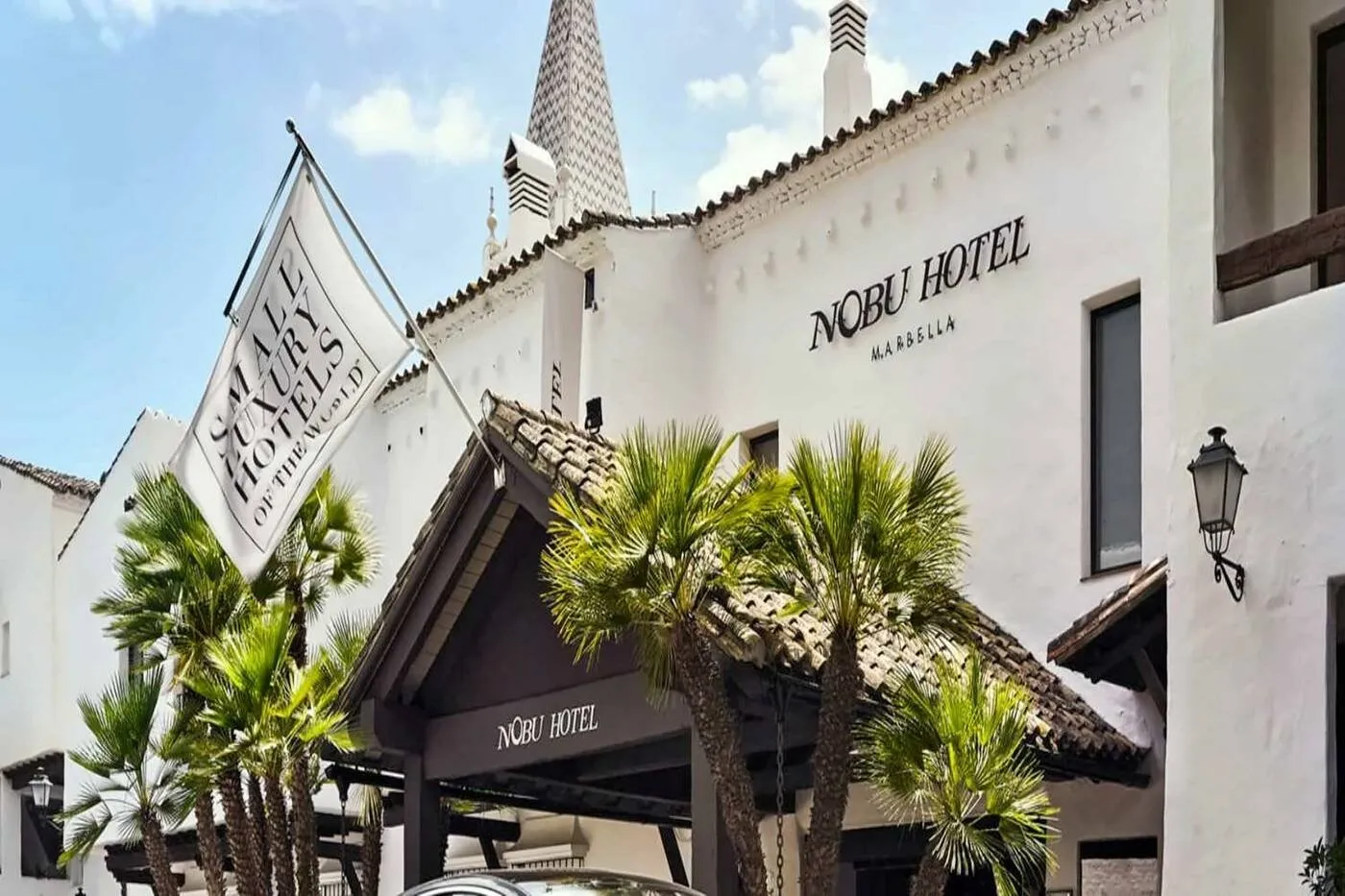 The entrance of Nobu Hotel Marbella: a tiled porch among fan palms below a whitewashed façade bearing the hotel's name