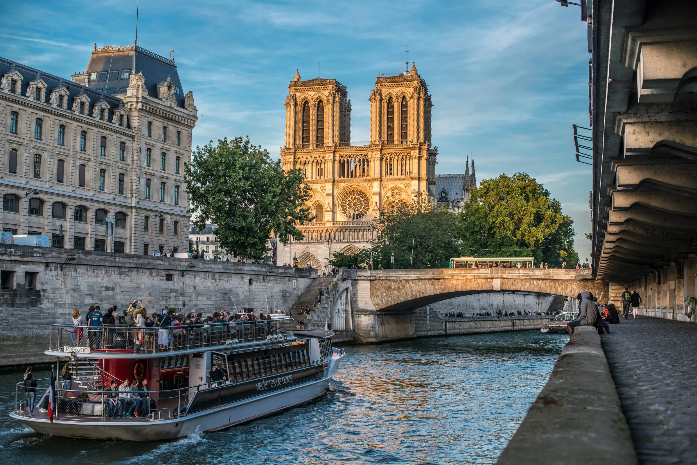 The west front of Notre-Dame in evening sun seen from the Left Bank quay, a sightseeing boat passing on the Seine below it, the Petit Pont and the island's quay buildings on the left