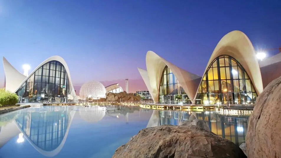 The Oceanogràfic in Valencia at dusk: white shell-shaped roofs over glass walls, mirrored in a still pool, with a white geodesic dome behind
