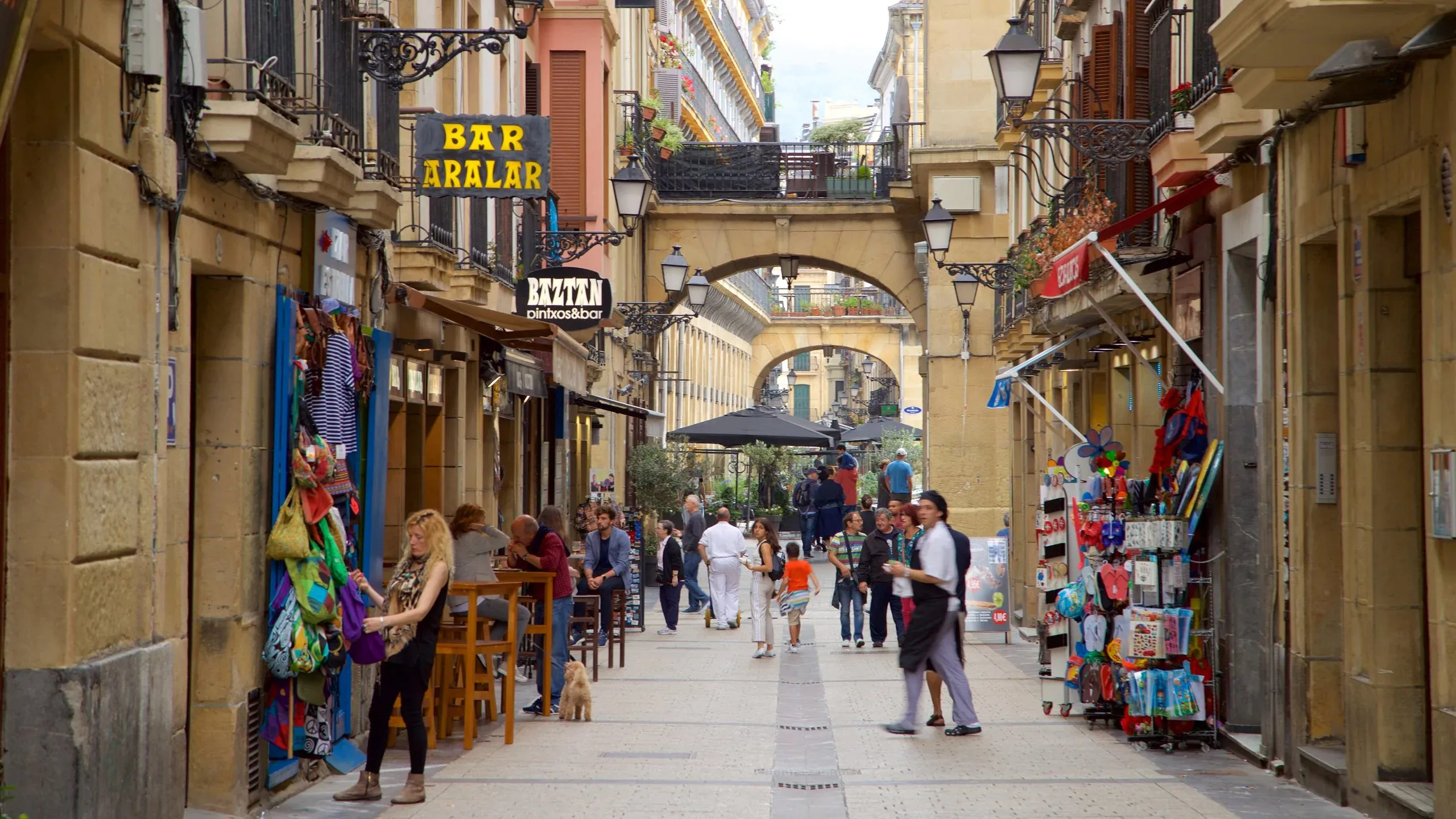 A pedestrian street in the Parte Vieja running under stone arches that bridge it between the houses, with pintxo bar signs and wrought-iron lamps on the walls, high tables outside a bar and people walking