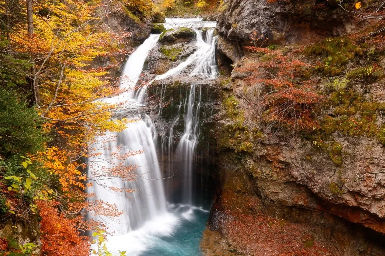 A waterfall dropping in white ribbons through beech woods in orange and gold into a turquoise pool, between mossy rock walls