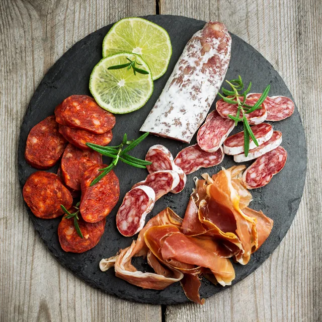 Cured meats on a round slate seen from above: rounds of chorizo and salchichón, a whole cured sausage, folds of cured ham and sprigs of rosemary, with two slices of lime