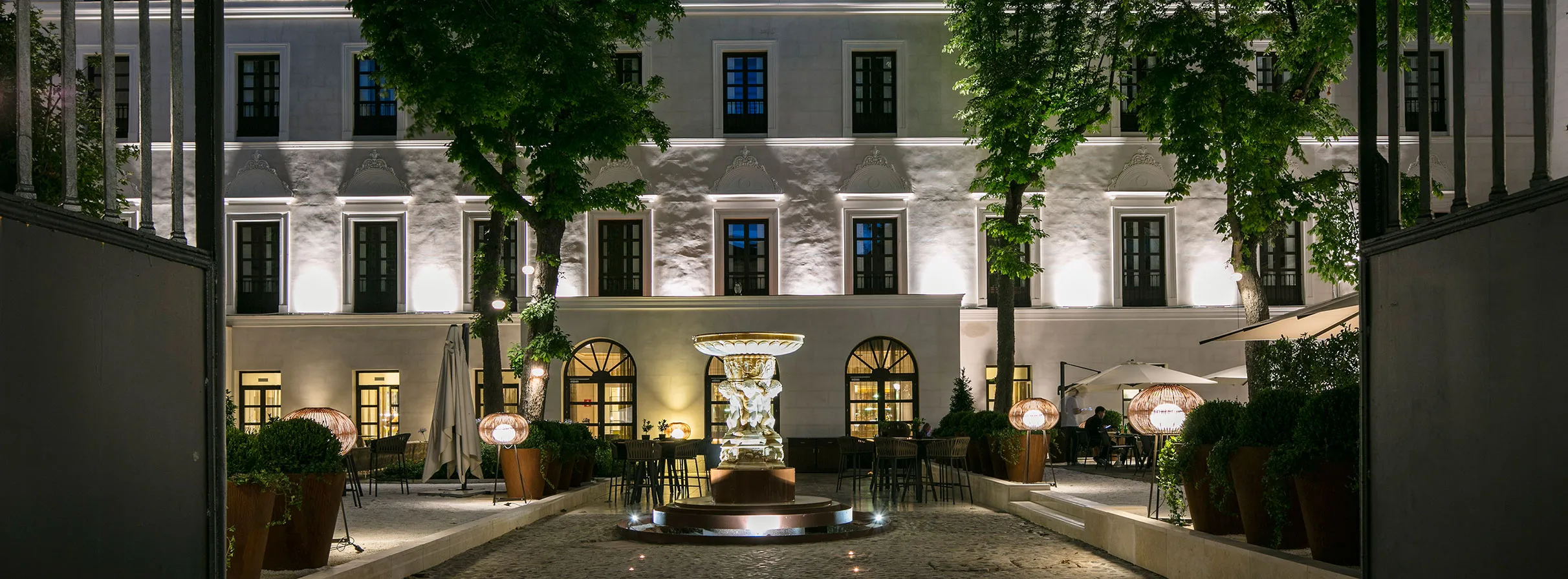The white façade of Palacio de los Duques lit at night, beyond a stone fountain and tables under the trees
