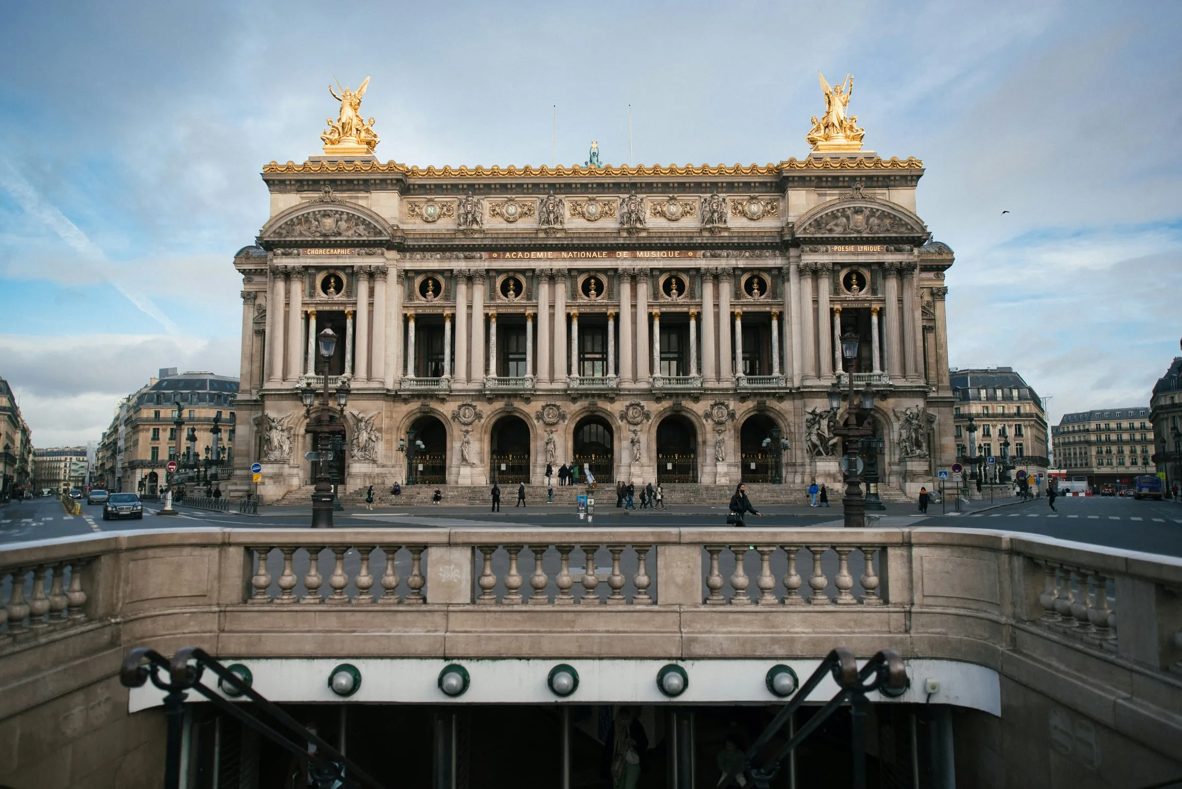 The Palais Garnier seen across the Place de l'Opéra from the top of the Métro steps: the columned façade with its gilded statues against a pale blue sky, Haussmann buildings on either side, lamp posts on the square and a stone balustrade in the foreground