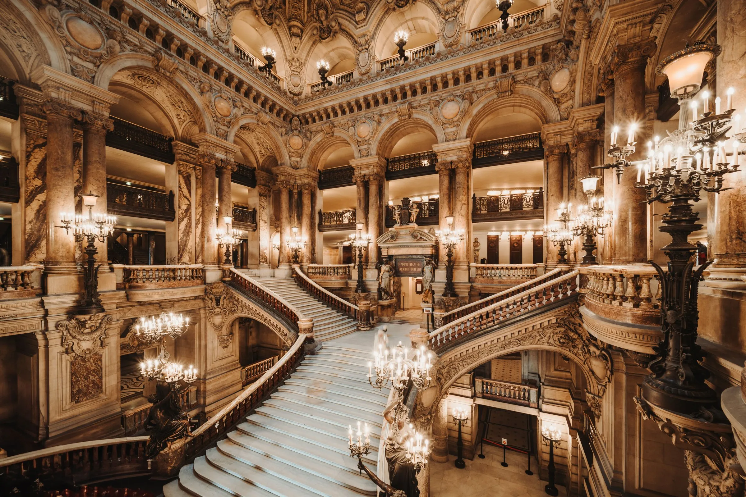 The grand staircase of the Palais Garnier: the double marble stair dividing under coloured marble columns and tiers of galleries, bronze figures holding candelabra at its foot, chandeliers lit along the balustrades and no one on the steps