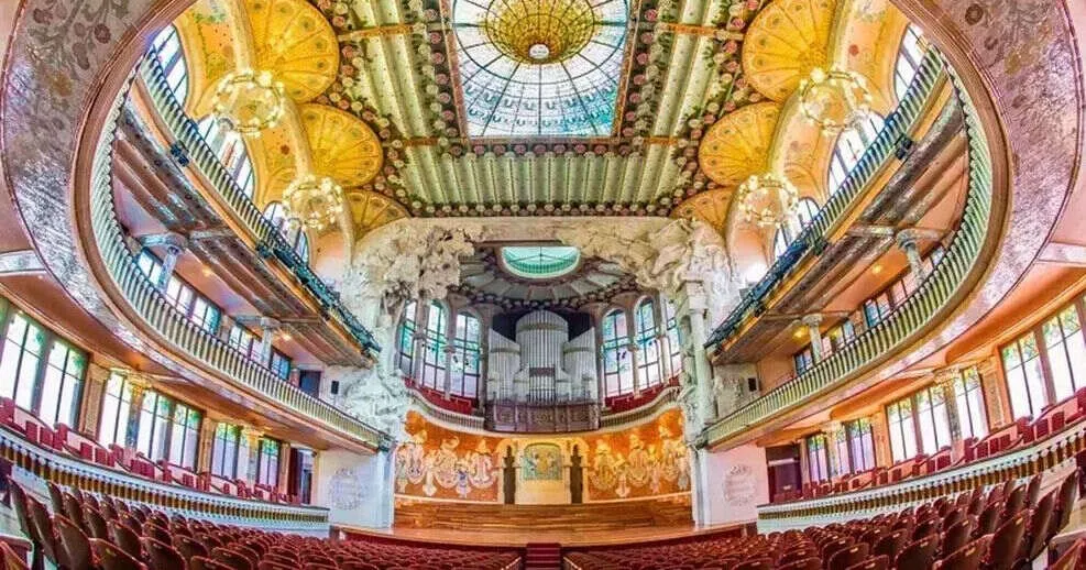 The concert hall of the Palau de la Música Catalana: a stained-glass skylight and painted ceiling over tiers of balconies, organ pipes behind a sculpted stage arch, and rows of red seats