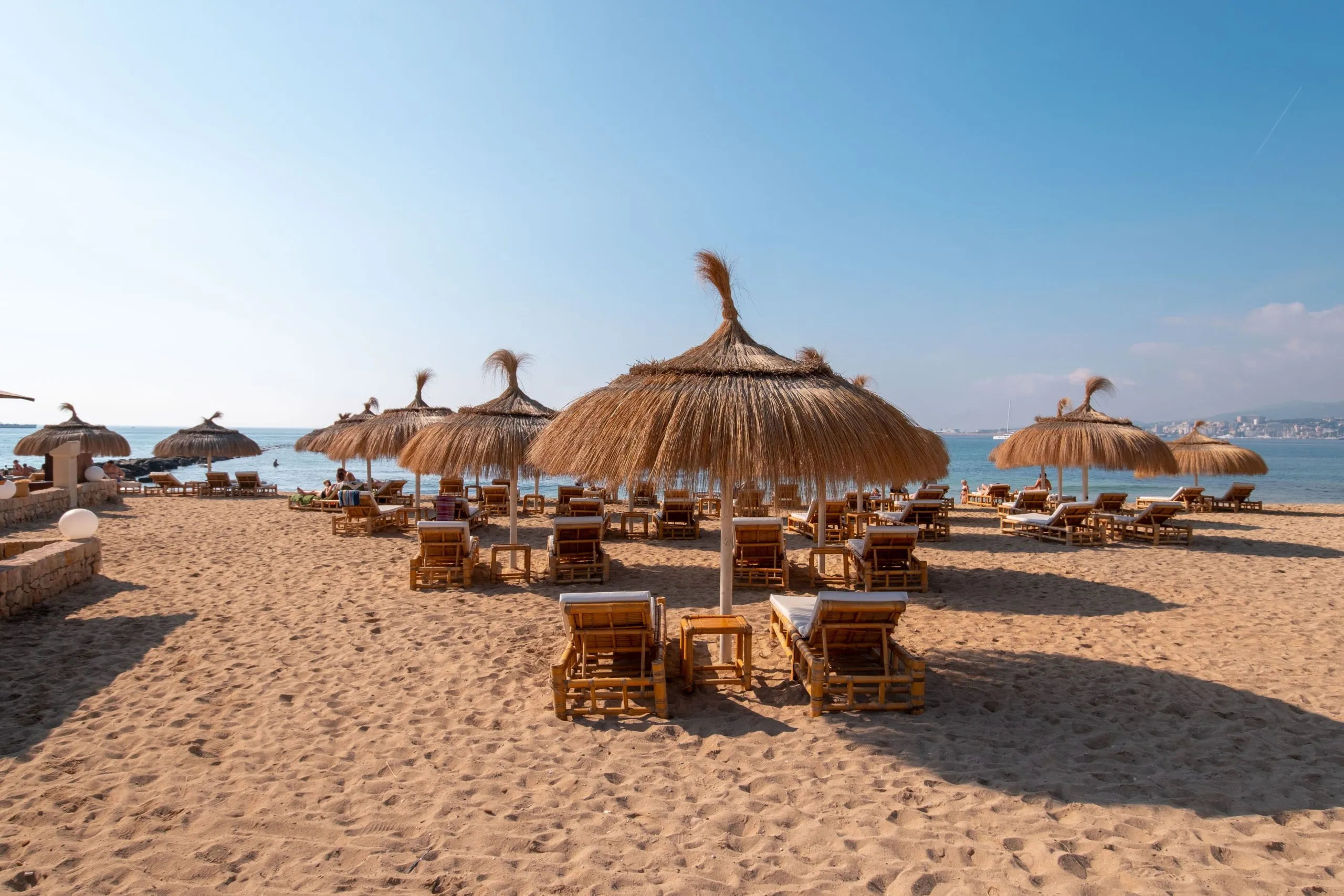 A beach club on the Bay of Palma: thatched parasols and bamboo loungers on raked sand, the turquoise bay and the city beyond