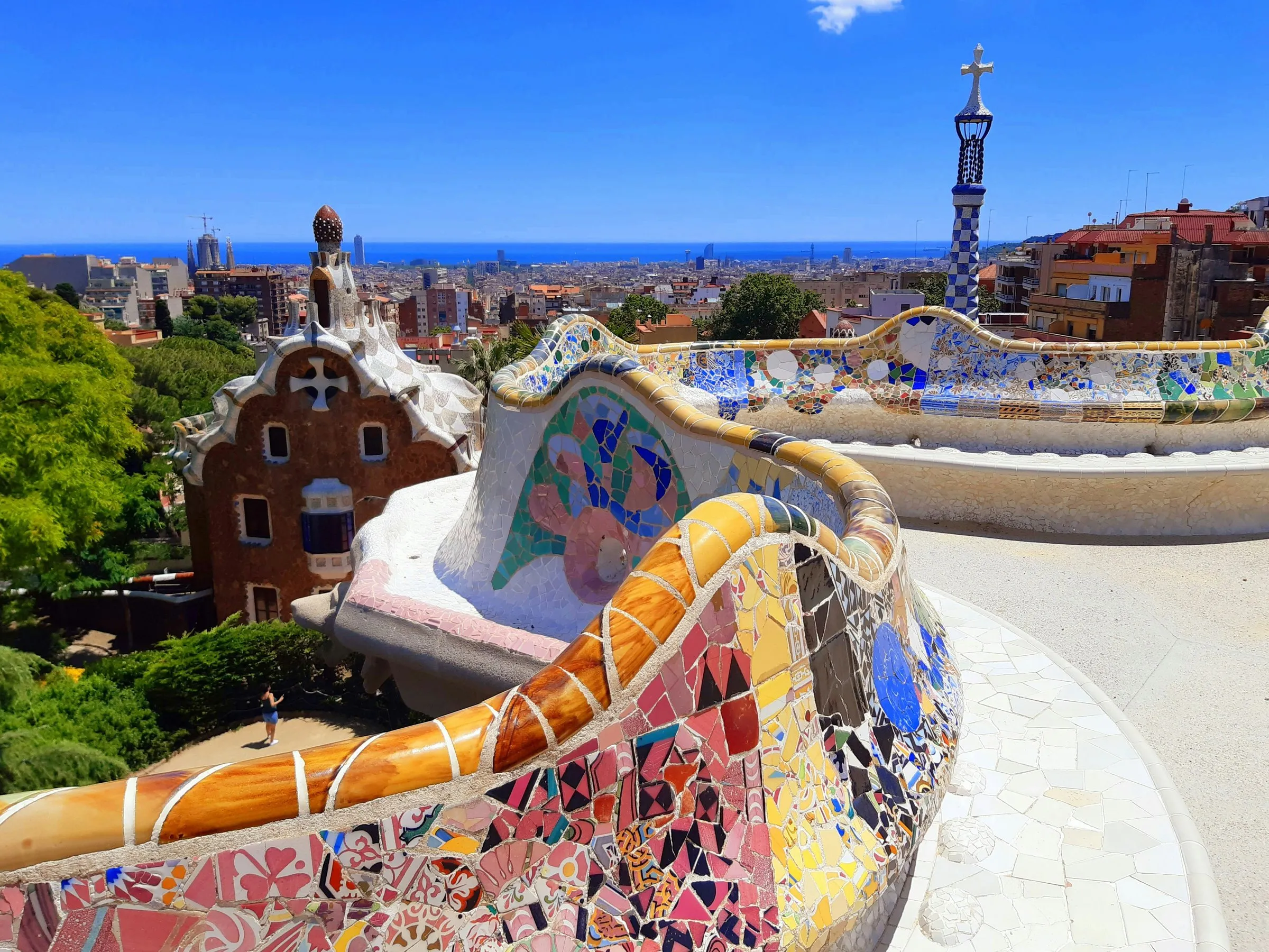 The serpentine trencadís bench of Park Güell curving through the foreground, one of Gaudí's gatehouses below it, and Barcelona running down to the sea under a clear blue sky