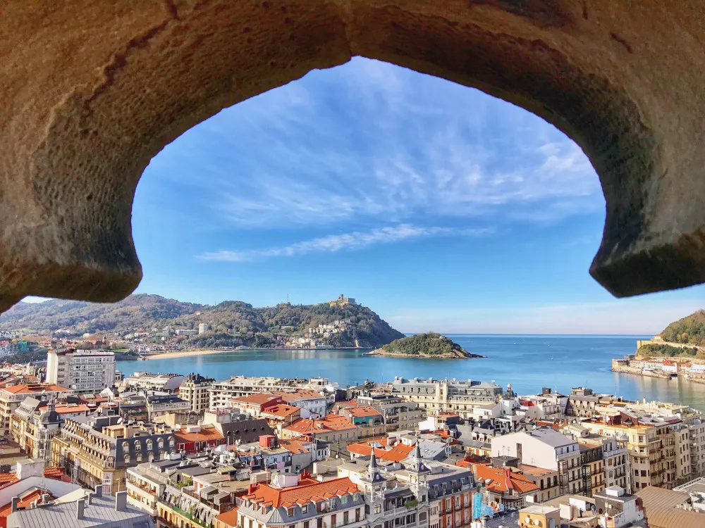 La Concha Bay framed by a rough stone arch high above the city's rooftops: Santa Clara island in the bay, Monte Igeldo on the far shore and the wooded slope of Monte Urgull at the right