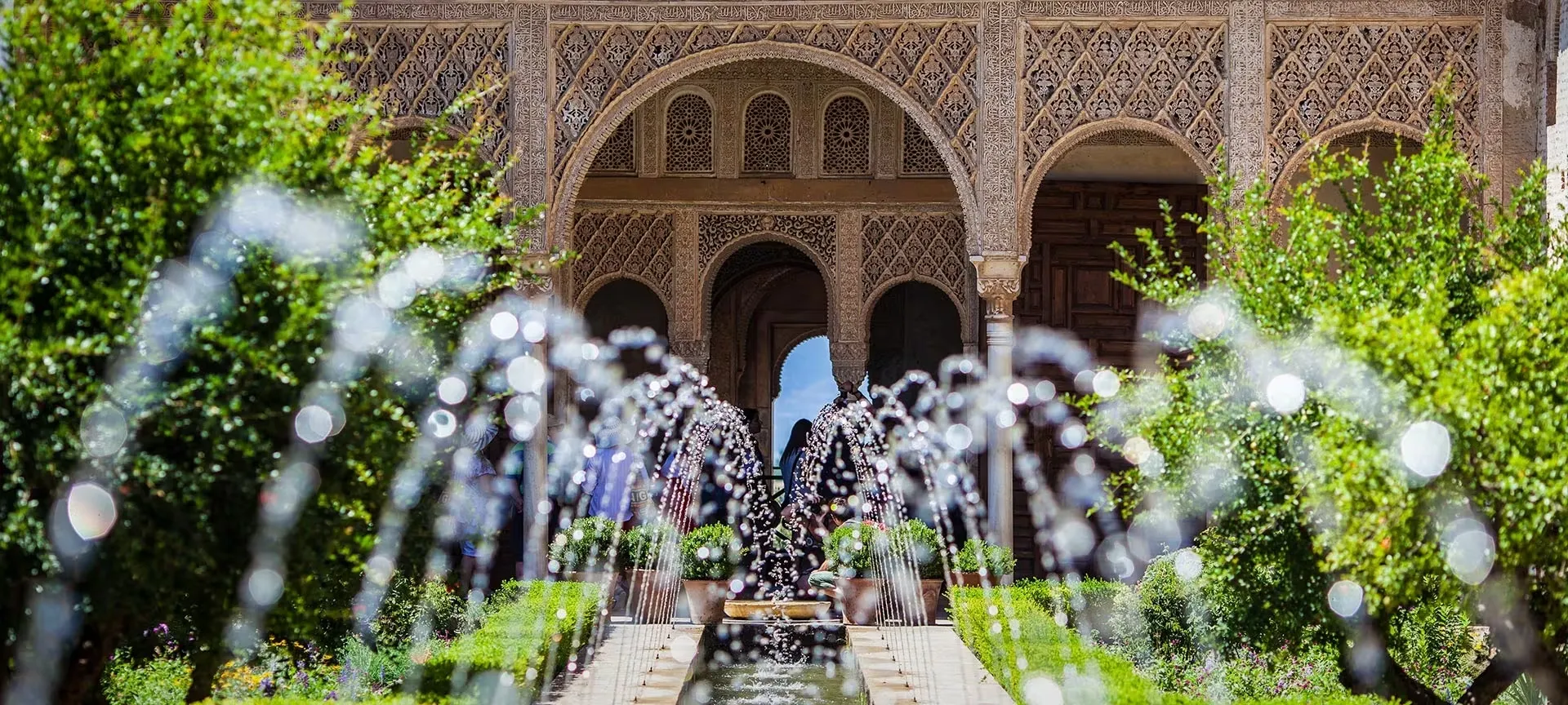 Fountain jets arching over a long, narrow pool between clipped shrubs and flowers, towards an arcaded pavilion of carved stucco