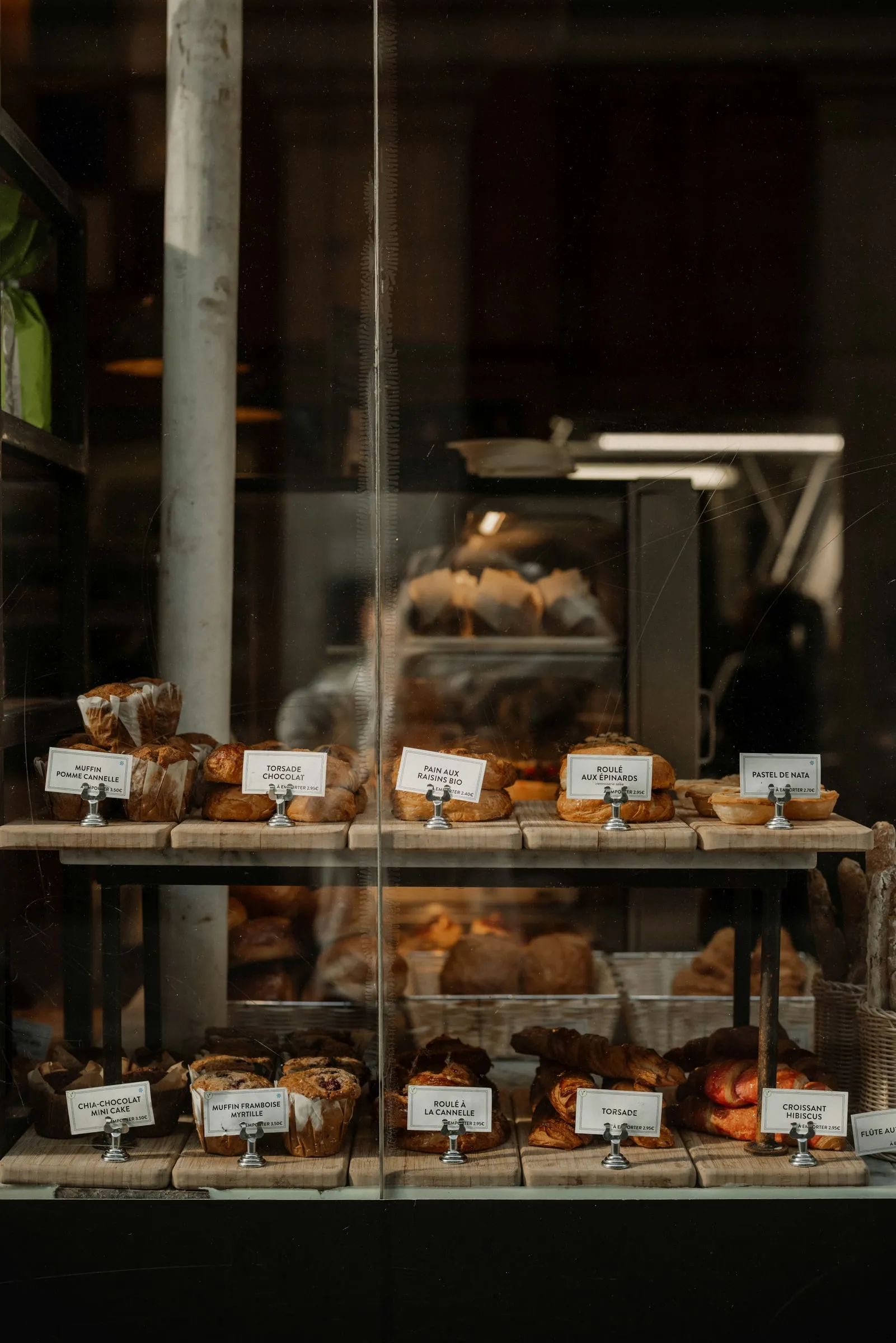Two wooden shelves of pastries behind a pâtisserie window, each with a small white card naming it in French — torsade, pain aux raisins, croissant — and the dark workshop out of focus behind