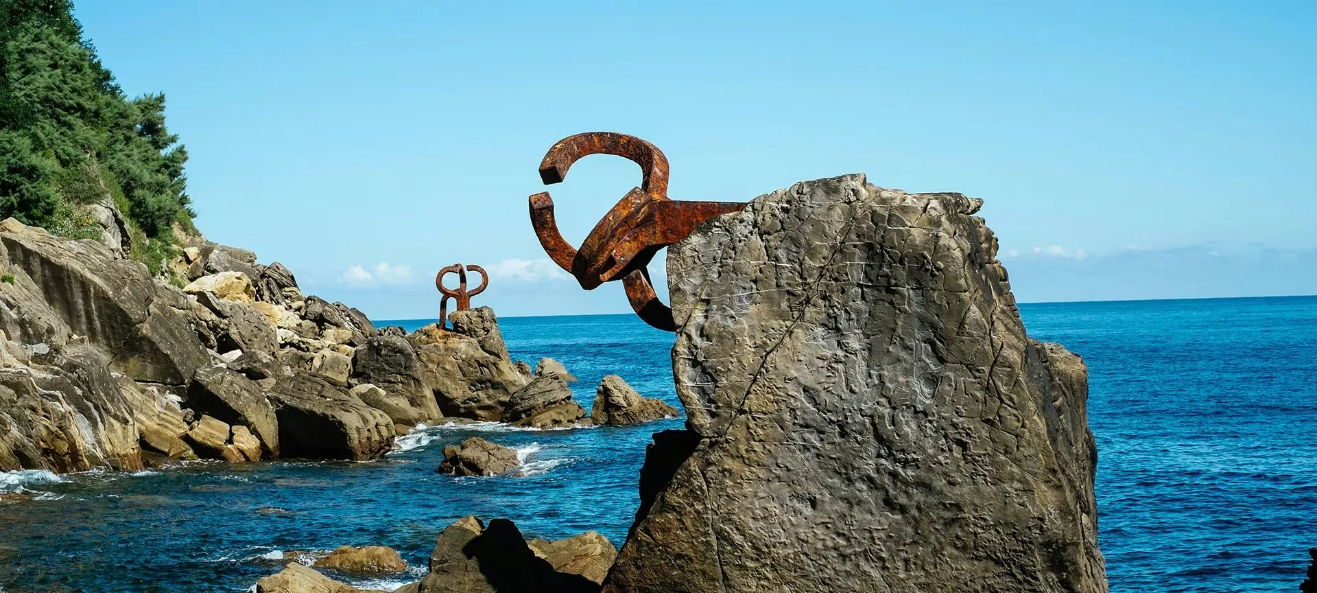 Rusted steel sculptures anchored to the rocks at the edge of a blue sea, one reaching out from a boulder in the foreground and a second on the rocks beyond