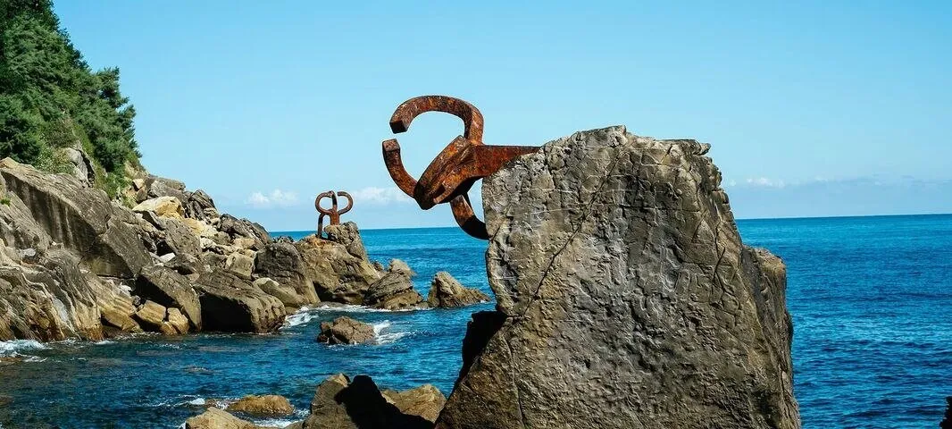 Rusted steel sculptures anchored to the rocks at the edge of a blue sea, one reaching out from a boulder in the foreground and a second on the rocks beyond