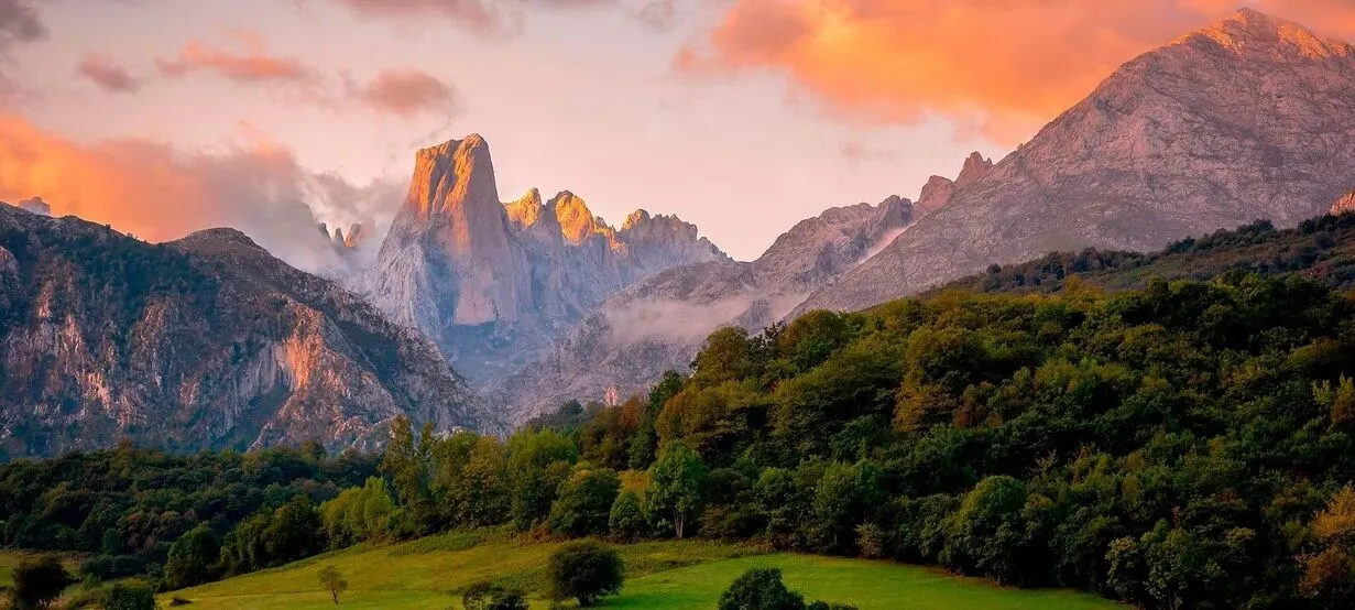The pale limestone tower of the Naranjo de Bulnes catching the sunset among grey peaks, under pink and orange cloud, above wooded hills and a green meadow