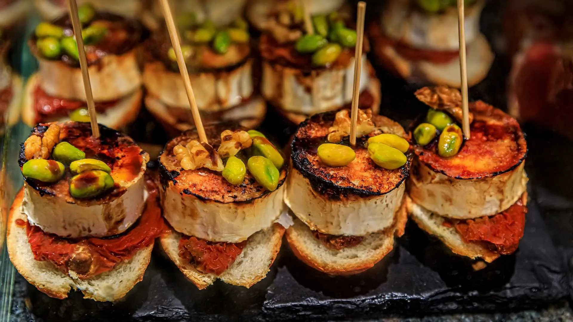 Pintxos on a slate: rounds of grilled goat's cheese with blackened tops on slices of bread and sun-dried tomato, scattered with pistachios and walnuts, each held by a wooden skewer