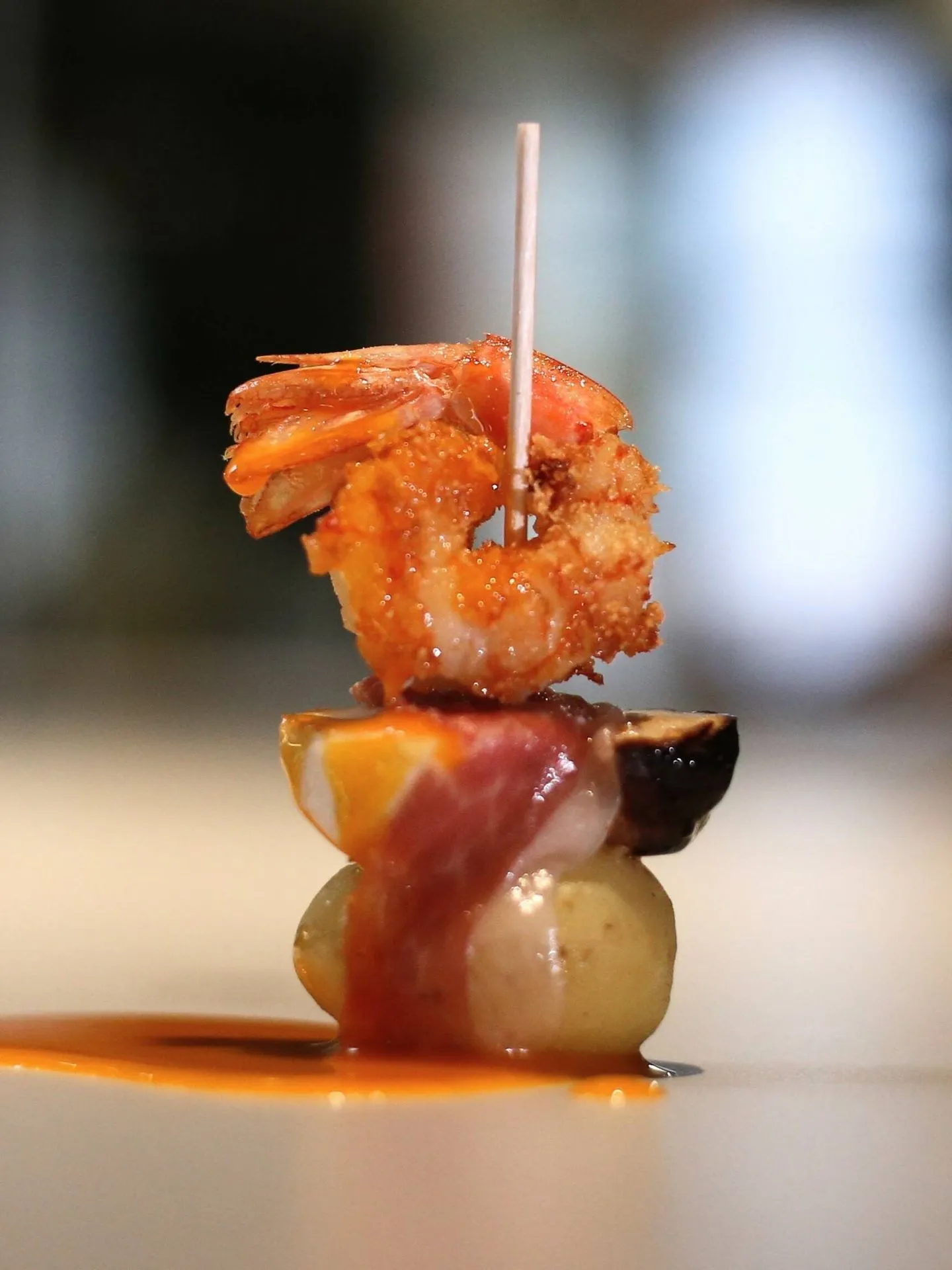 A pintxo on a cocktail stick: a crisp fried prawn on top of a small potato wrapped in cured ham, standing in a spill of orange sauce on a pale counter