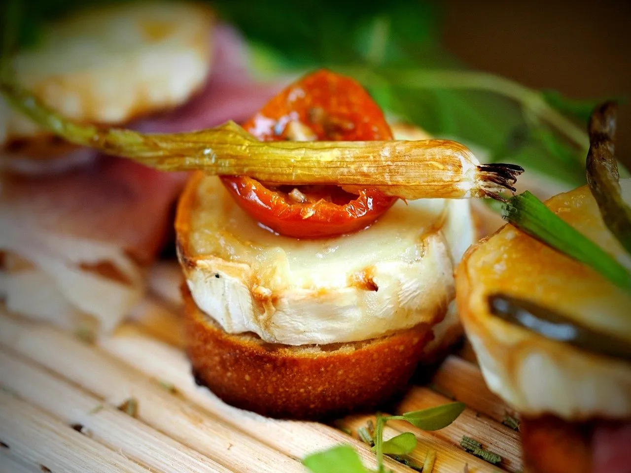 A close-up of a pintxo on toasted bread: melted goat's cheese topped with half a roasted cherry tomato and a charred spring onion, more pintxos blurred beside it on a wooden board