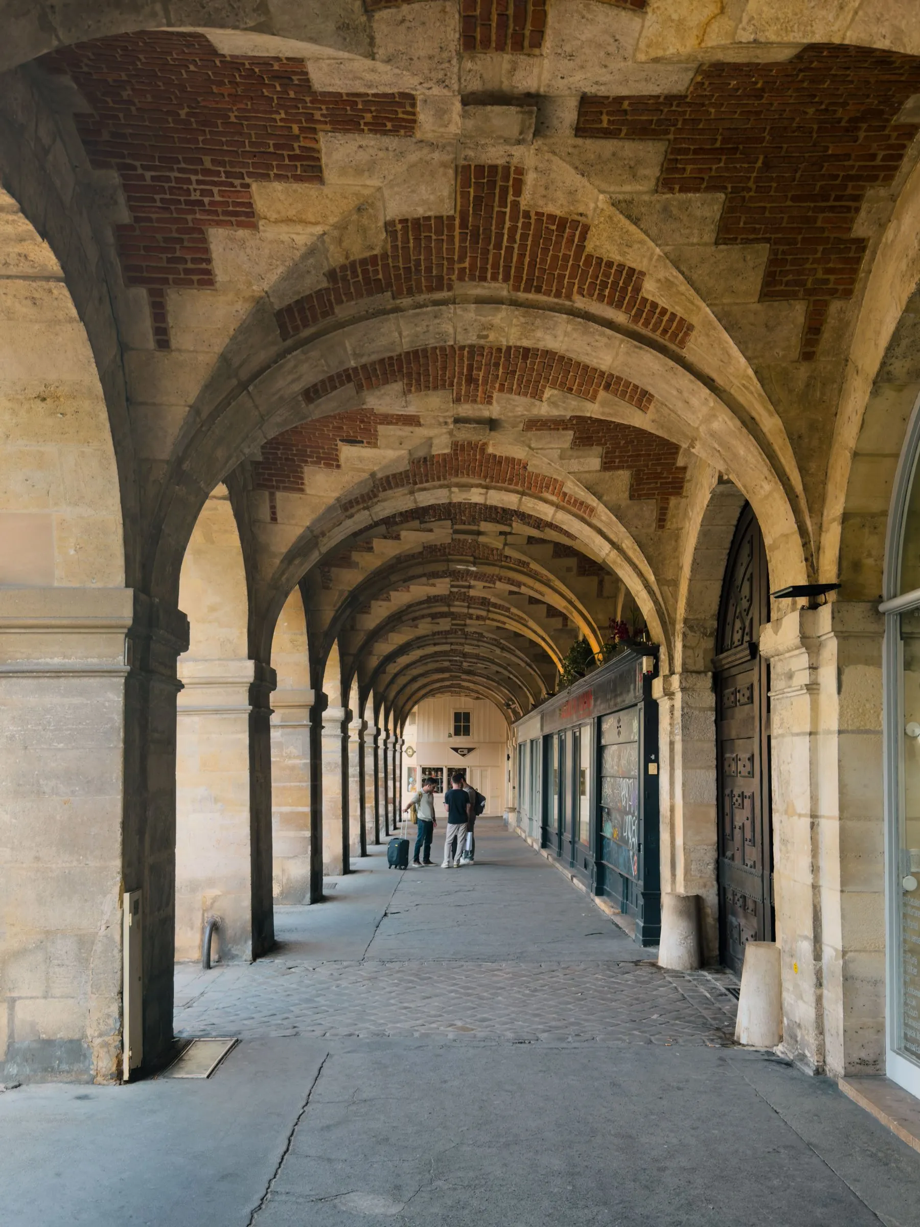 The arcade of the Place des Vosges: a long run of stone arches with groin vaults infilled in red brick, shopfronts and a studded wooden door beneath them on the right, and two visitors with a suitcase small in the distance where the arcade opens to daylight