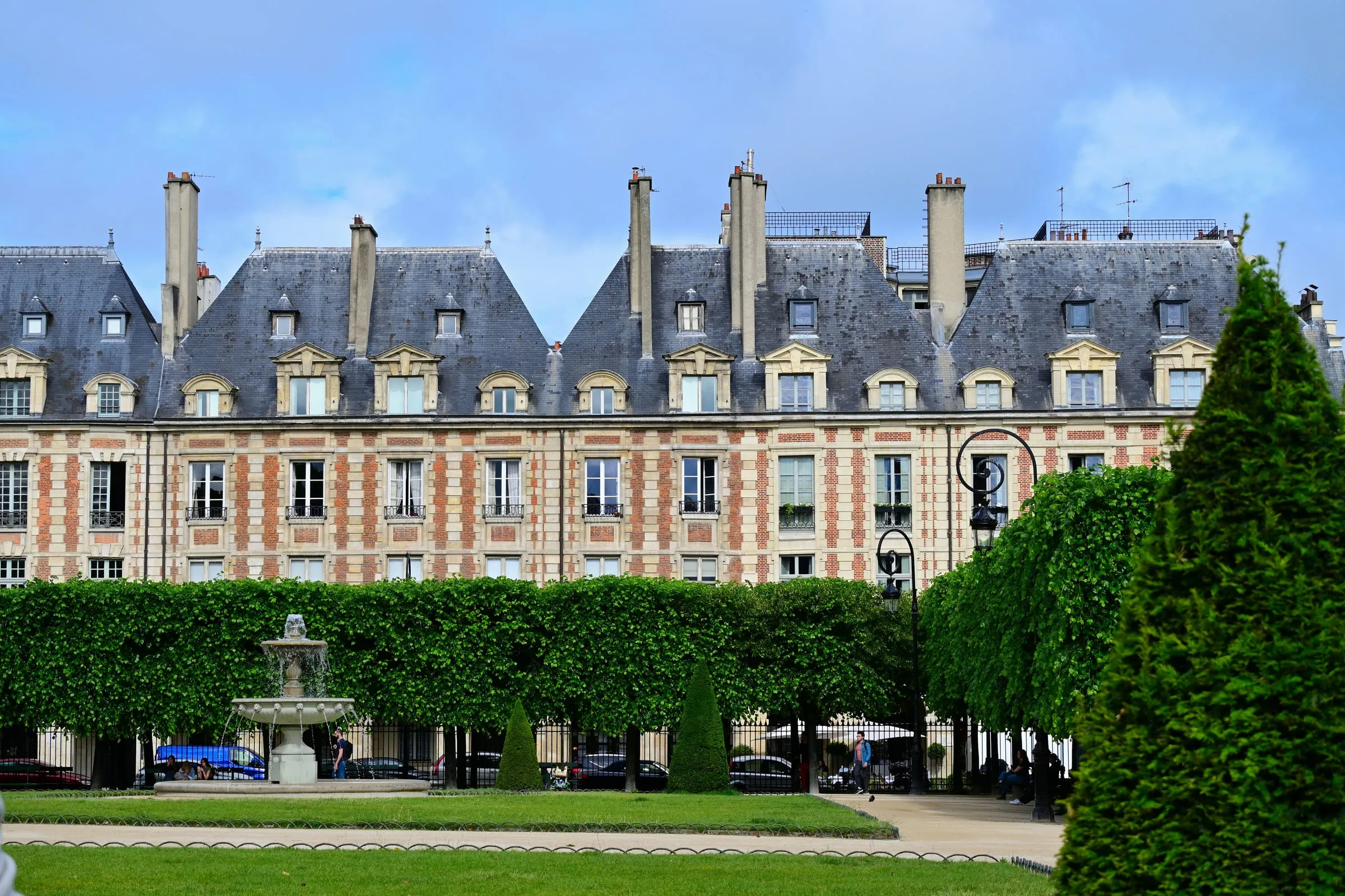 The red-brick and stone pavilions of the Place des Vosges with their steep slate roofs and dormers, a wall of clipped lime trees and a stone fountain in front of them, under a cloudy sky