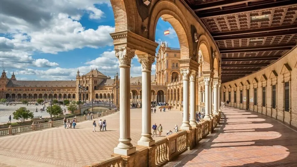 Looking out from the Plaza de España's arcade: white columns and brick arches frame the curving building, a tower flying the Spanish flag and a bridge over the canal, with people crossing the square