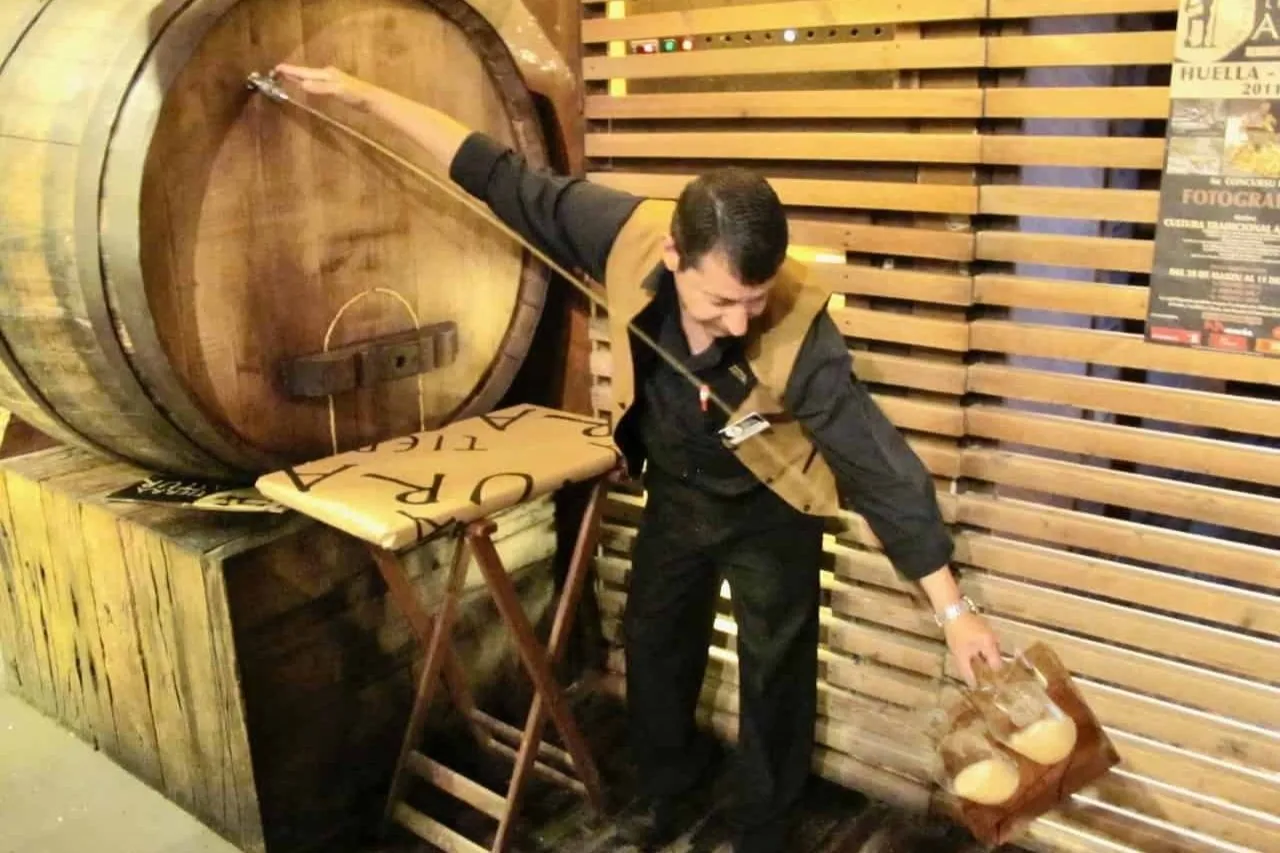 A waiter in a cider house catching cider from the tap of a great wooden barrel, his arm raised high to the tap and a glass held low at his side, against a wall of wooden slats