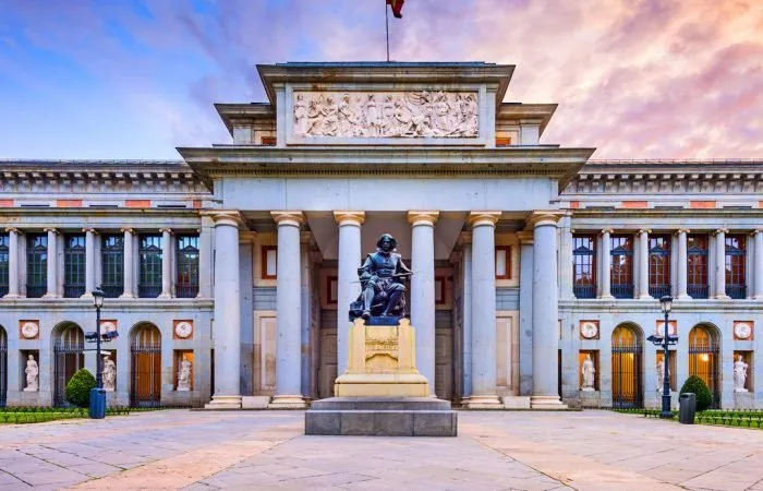 The Prado's columned entrance at dusk, with a bronze statue of the seated painter Velázquez on a stone pedestal before it and a pink and blue sky above