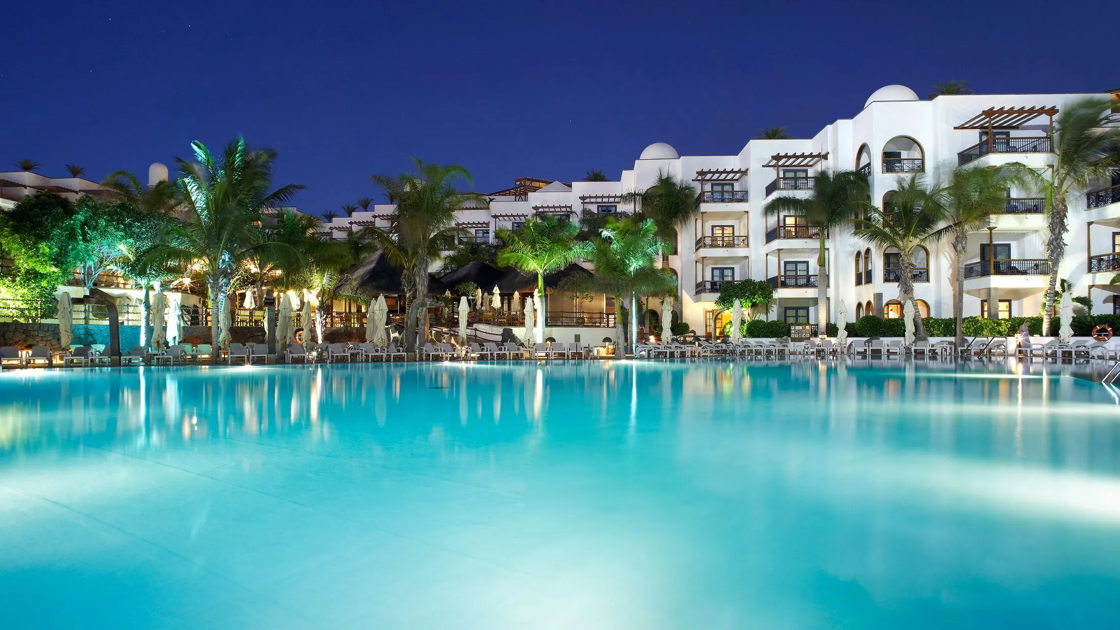 The main pool at Princesa Yaiza at night, the lit white arcades and palms of the resort reflected in still water
