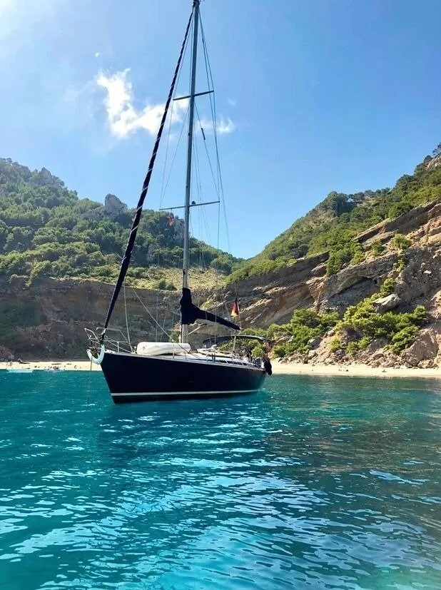 A dark-hulled sailing yacht at anchor in clear turquoise water off a sandy beach, beneath steep pine-covered cliffs