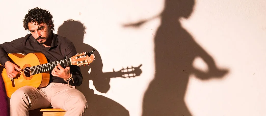 A flamenco guitarist playing on a low stool against a sunlit white wall, his shadow beside him and, further along the wall, the shadow of a dancer with her arms raised