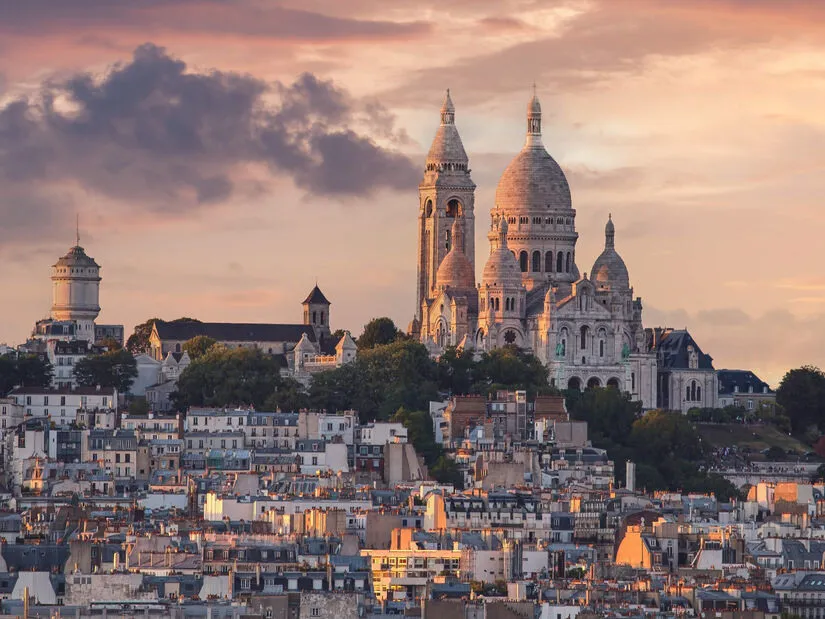 The white domes of the Sacré-Cœur on the hill of Montmartre at sunset, above trees and a slope of Paris rooftops, under pink clouds
