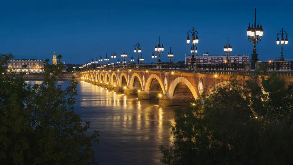 The Pont de Pierre in Bordeaux at dusk, its brick arches and street lamps lit gold above the wide Garonne, with the stone quay and a church spire on the far bank under a deep blue sky