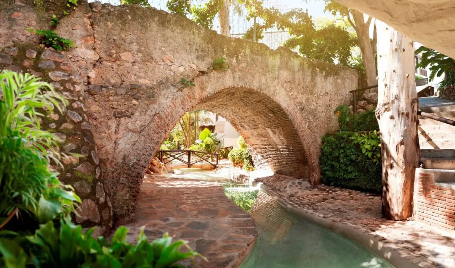 A single arch of rough stone and brick spanning a cobbled path and a shallow stream, with ferns and palms in the foreground, a pale tree trunk at the right and steps climbing beside it