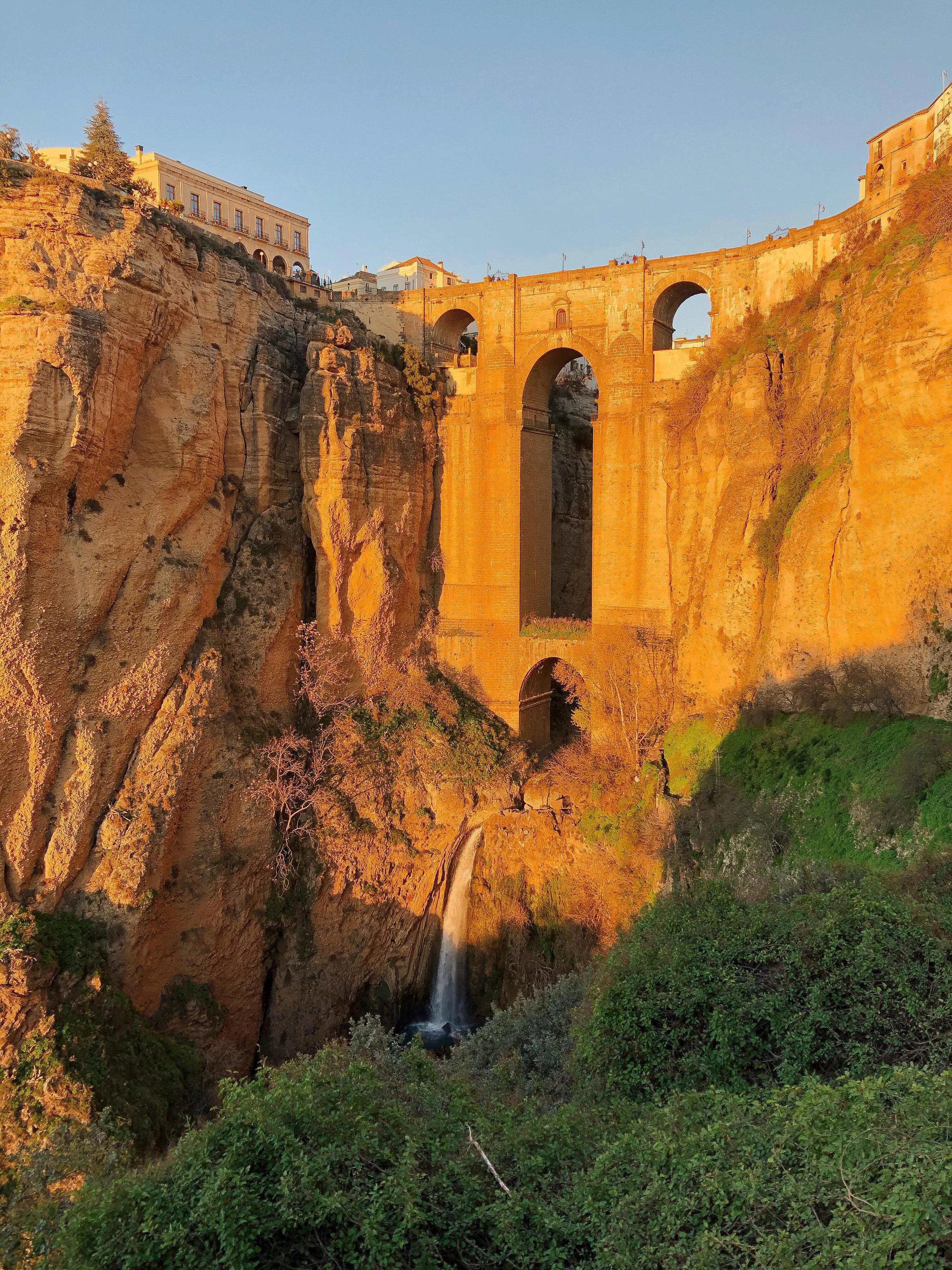 The Puente Nuevo spanning a deep gorge between cliffs lit gold by a low sun, a thin waterfall falling below its arches, green scrub in the foreground and buildings along both clifftops
